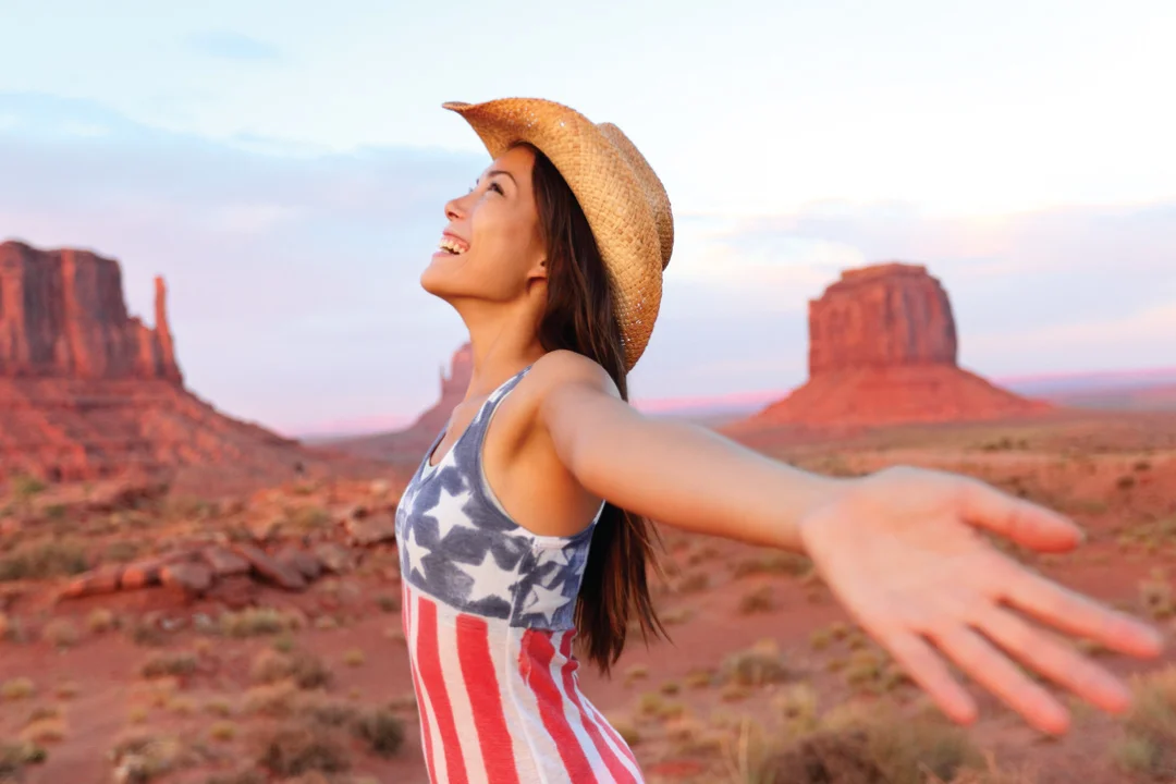 young female adult wearing cowboy hat and patriotic tank top with her arms to the wind, smiling and looking at sunset