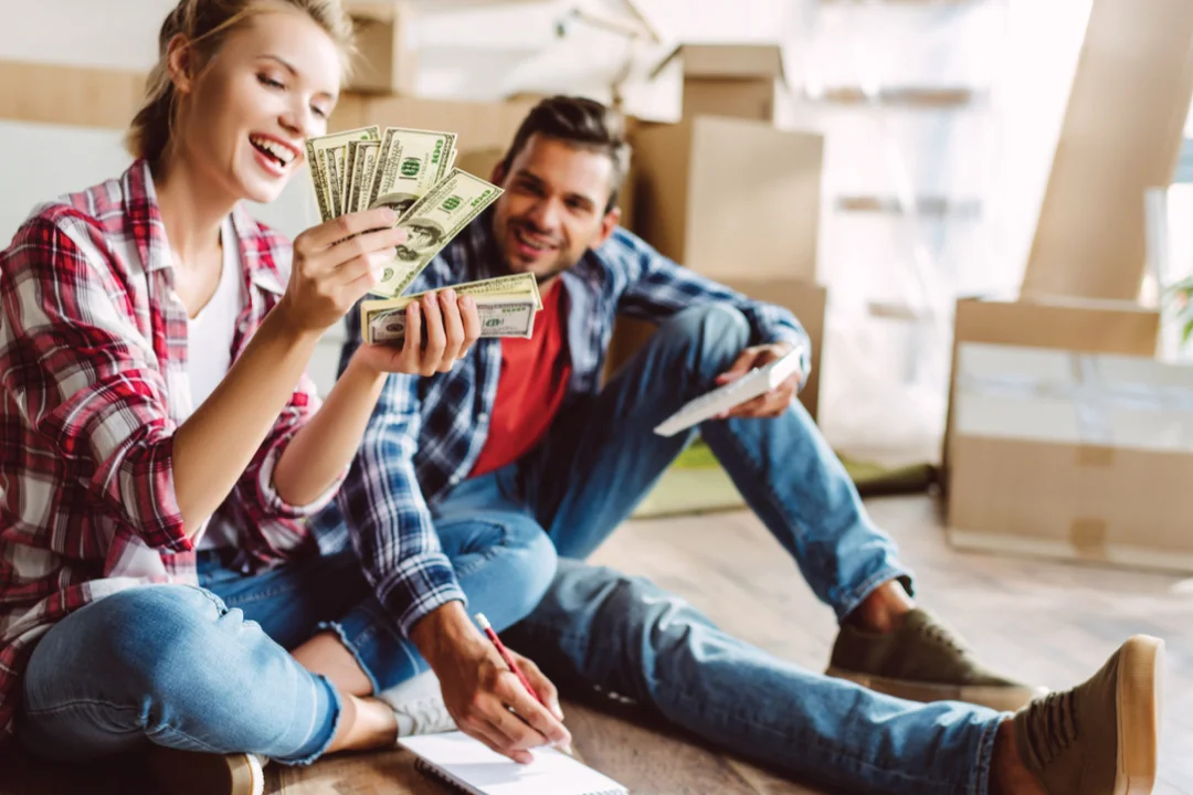 young couple sitting on ground of new house with moving boxes behind them, couple sitting and counting 100 dollar bills